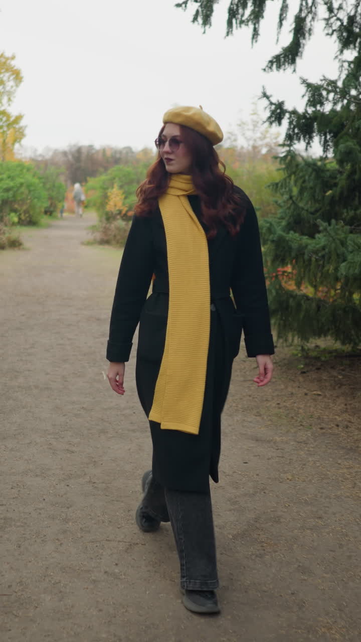 Giovane donna con lunghi capelli rossi ondulati che indossa un basco giallo, una sciarpa e occhiali da sole, guardandosi intorno felicemente con una vista sfocata di due persone sullo sfondo lontano.