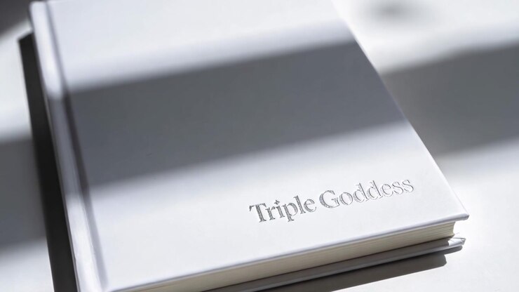 Sliding blind shadows across white book on desk, camera zooming revealing raised Triple Goddess
