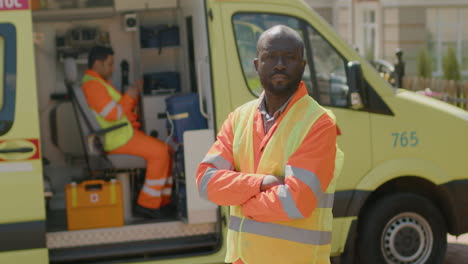 medical assistance with crossed arms standing on the street while his colleague is inside the ambulance