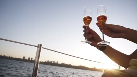 Two-crystal-glasses-with-wine-or-champagne-on-background-of-sea,-sunlight-and-blue-sky.-Man-and-woman-are-holding-their-and-clinking-by-them,-luxury-celebration-on-the-yacht