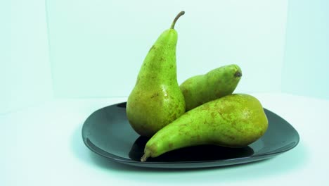 fresh big green pears rotates slowly on a black plate on light blue background, healthy food concept, medium shot, camera rotate left