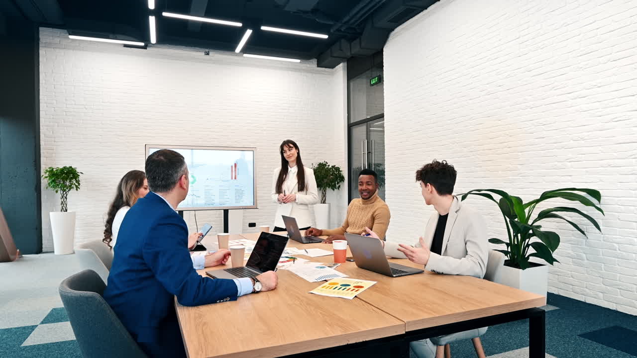 Jefa de equipo en una reunión de negocios en una oficina, discutiendo asuntos de negocios con los trabajadores utilizando una gran pantalla con gráficos, papeles y dispositivos en la mesa