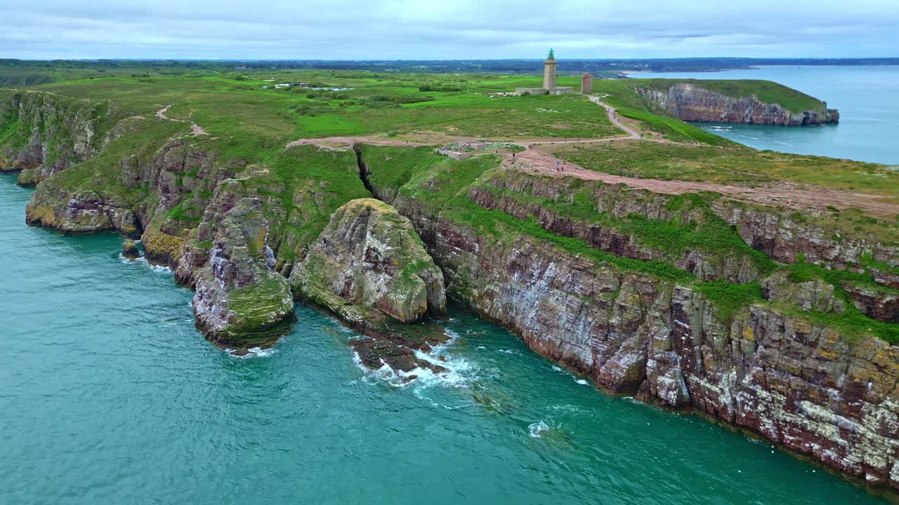 Drone pulls away from Cap Fréhel cliffs, revealing rugged coast, green trails, and lighthouse in Brittany, France.