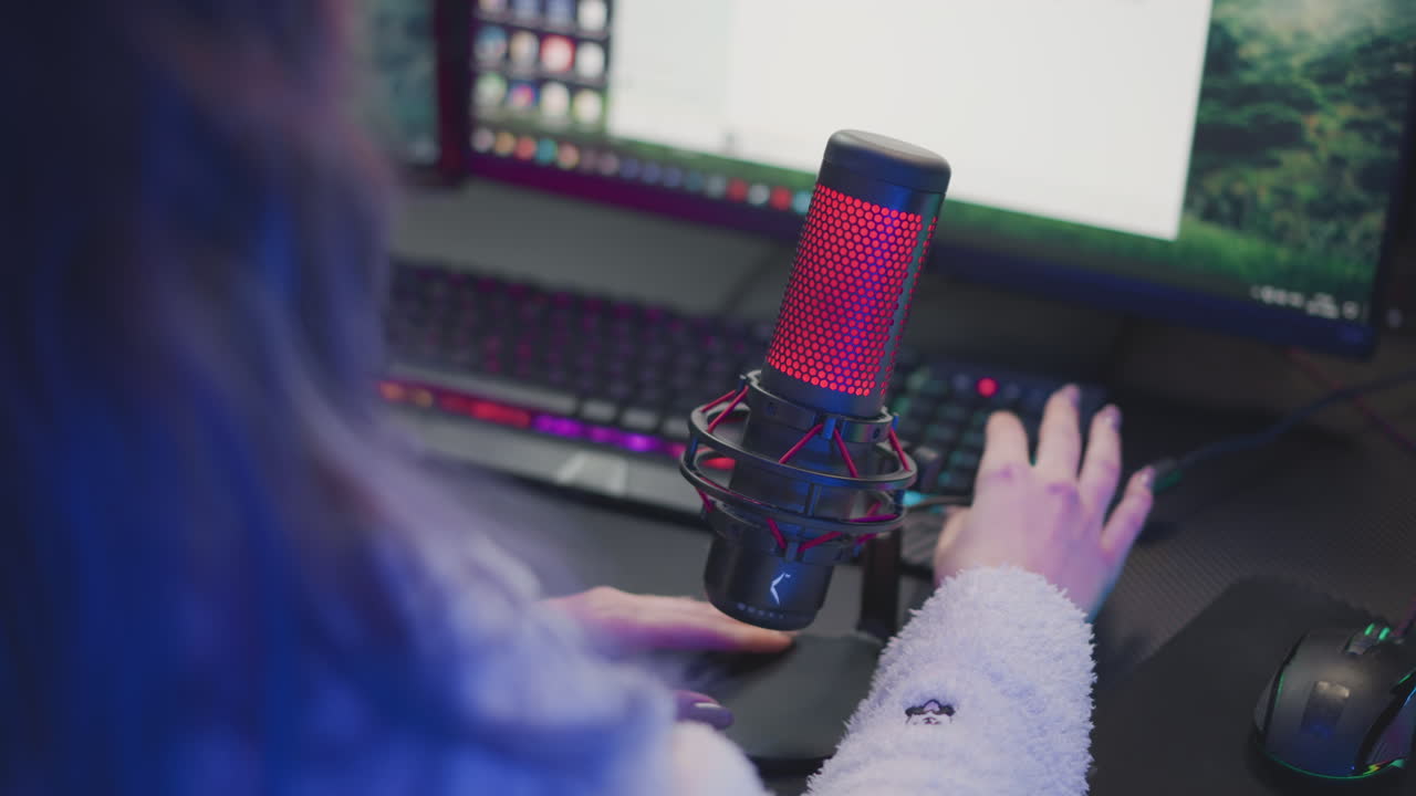 rear view of lady in fluffy jacket bringing red studio microphone forward and adjusting cable beside rgb light strip keyboard monitor for streaming podcast recording setup in cozy high tech workspace