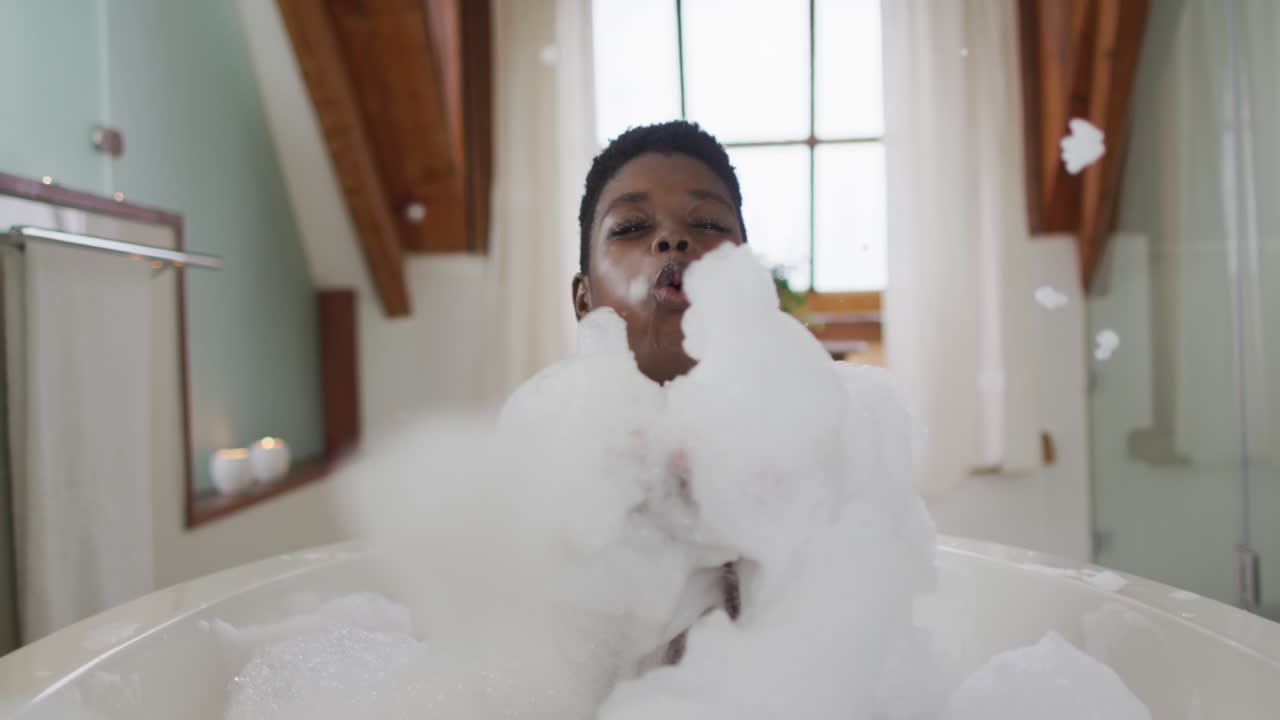 Smiling african american attractive woman taking bath and blowing out foam in bathroom