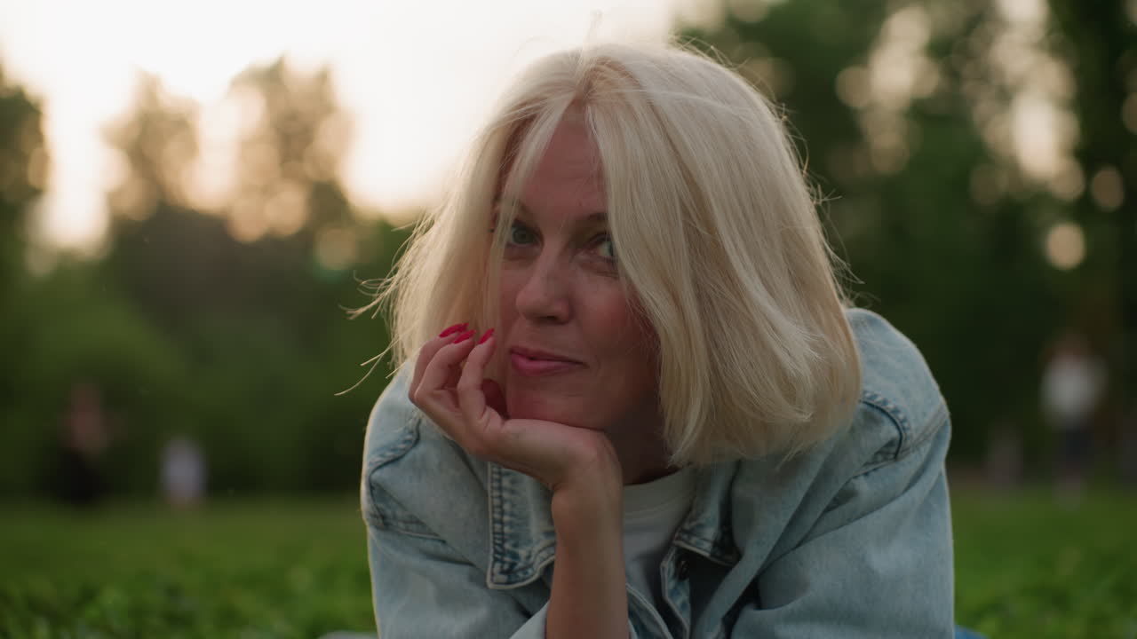 Ritratto di mamma premurosa sdraiata su una coperta da picnic al parco che usa la mano per sostenere la testa mentre sorride dolcemente, persone sfocate che giocano sullo sfondo, calda luce serale, atmosfera familiare rilassata