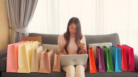 attractive asian girl surrounded with shopping bags using her credit card for shopping online, type in card information on the laptop