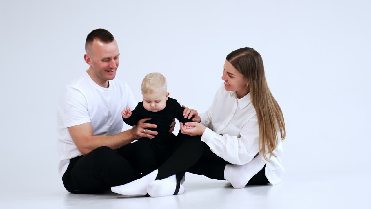 Beautiful Caucasian parents in white tops and black pants sit on the floor. Smiling mom and dad hold their adorable baby boy. White background.