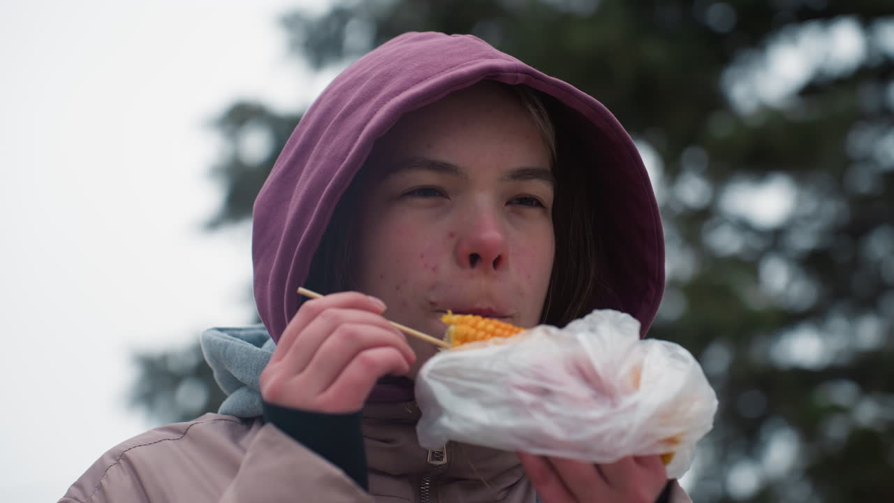 겨울 재을 입은 어린 소녀가 옥수수를 먹고, 거리 음식을 즐기고, 후드 재, 캐주얼 야외 간식, 겨울 설정, 막대기 음식, 추운 날씨에 먹고, 플라스틱으로 포장, 도시 배경