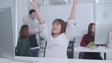 excited asian businesswoman sitting at her office desk arms raised celebration of successful project, job promotion.