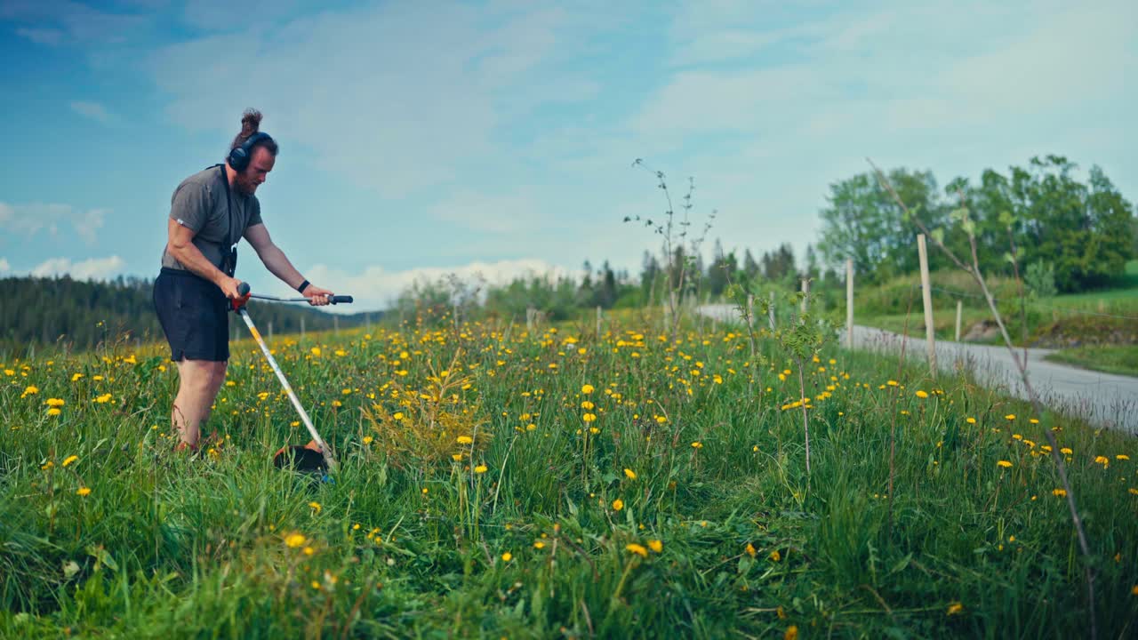 Caucasian Guy Using A Hand-held Lawn Mower Over Dense Grass. Wide Shot