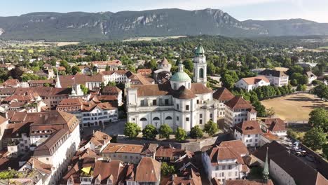 aerial view captures the city of olten, switzerland, highlighting a prominent, domed church in its center. the historic cityscape is framed by rolling green hills under a bright, clear sky, solothurn