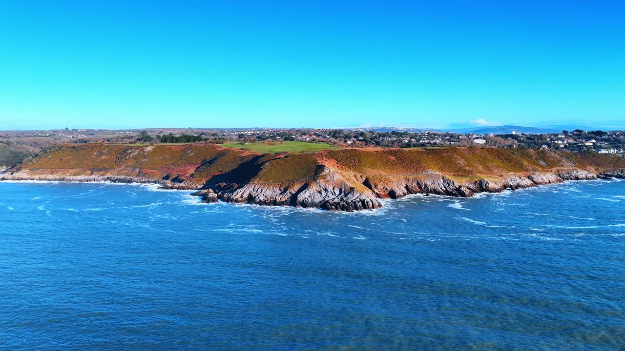 Panorama Aéreo do Litoral Galês com Cidade em Colinas Distantes. Um longo trecho de costa de falésias se estende em direção a uma cidade na encosta enquanto ondas quebrando traçam a costa rochosa sob céu claro