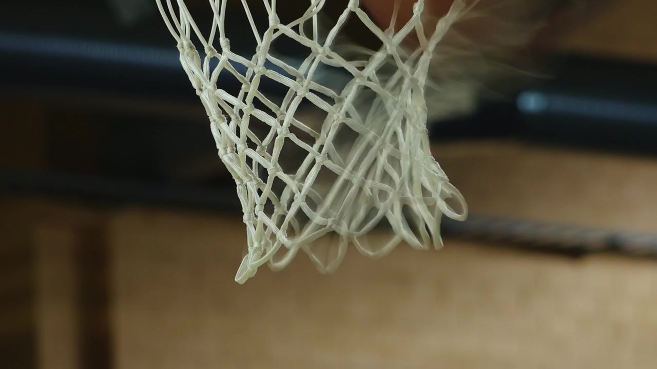 Primo piano di un pallone da basket che cade attraverso una rete bianca in un campo al coperto, con una leggera sfocatura di movimento e una messa a fuoco ridotta che enfatizzano l'azione e lo sport.