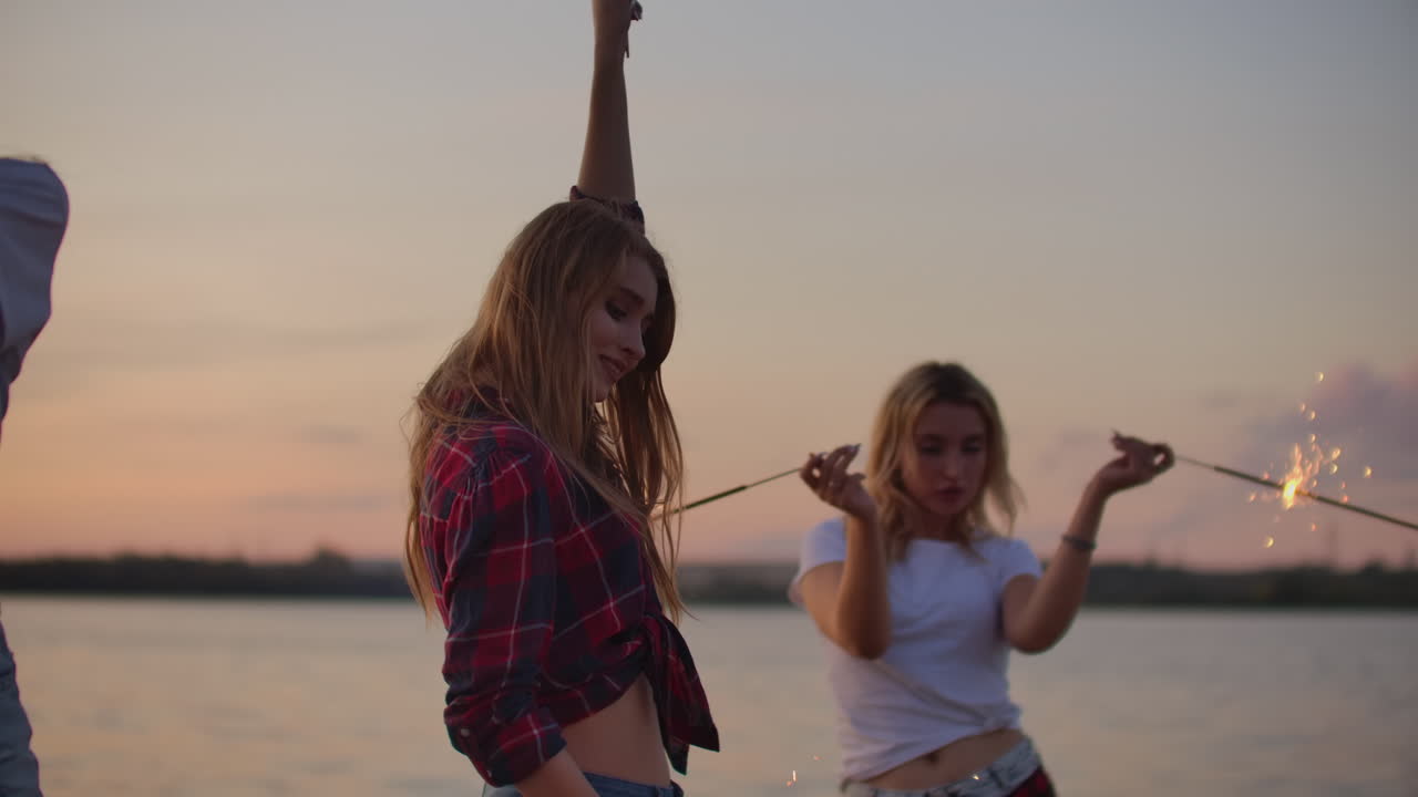 dos chicas están bailando con grandes luces bengalíes en la costa del río. esta es una noche de verano fresca en la fiesta al aire libre al atardecer rosado.