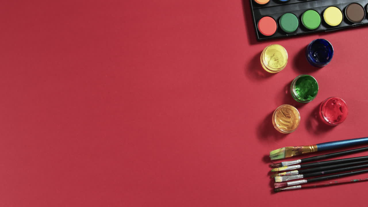 vista aérea de pinturas y pinceles dispuestos sobre un fondo rojo con espacio de copia, en cámara lenta