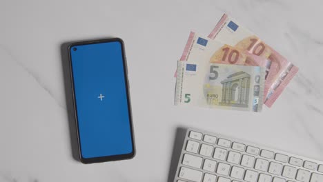 Toma-De-Moneda-Aérea-De-Mano-Escribiendo-En-El-Teclado-De-La-Computadora-Con-Billetes-De-Euro-Y-Teléfono-Móvil-De-Pantalla-Azul