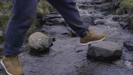 close-up of feet walking across a small river creek in brown hiking boots and dirty pants in nordic landscape in slow motion on the faroe islands