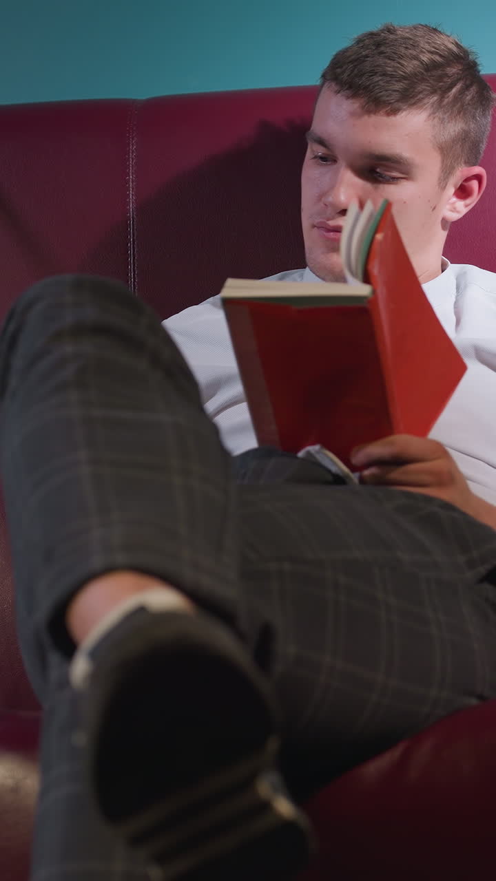 un tipo con camisa blanca y pantalones a cuadros se sienta en un sofá rojo en la sala de billar, con las piernas cruzadas, leyendo un libro mientras tira una pelota de billar naranja en la mano.