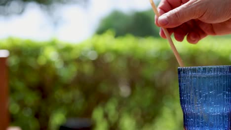 hand inserting straw into sparkling beverage outdoors with greenery background