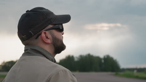 side view of male field technician in dark sunglasses and cap holding smartphone monitoring weather conditions beside rural road under cloudy sky
