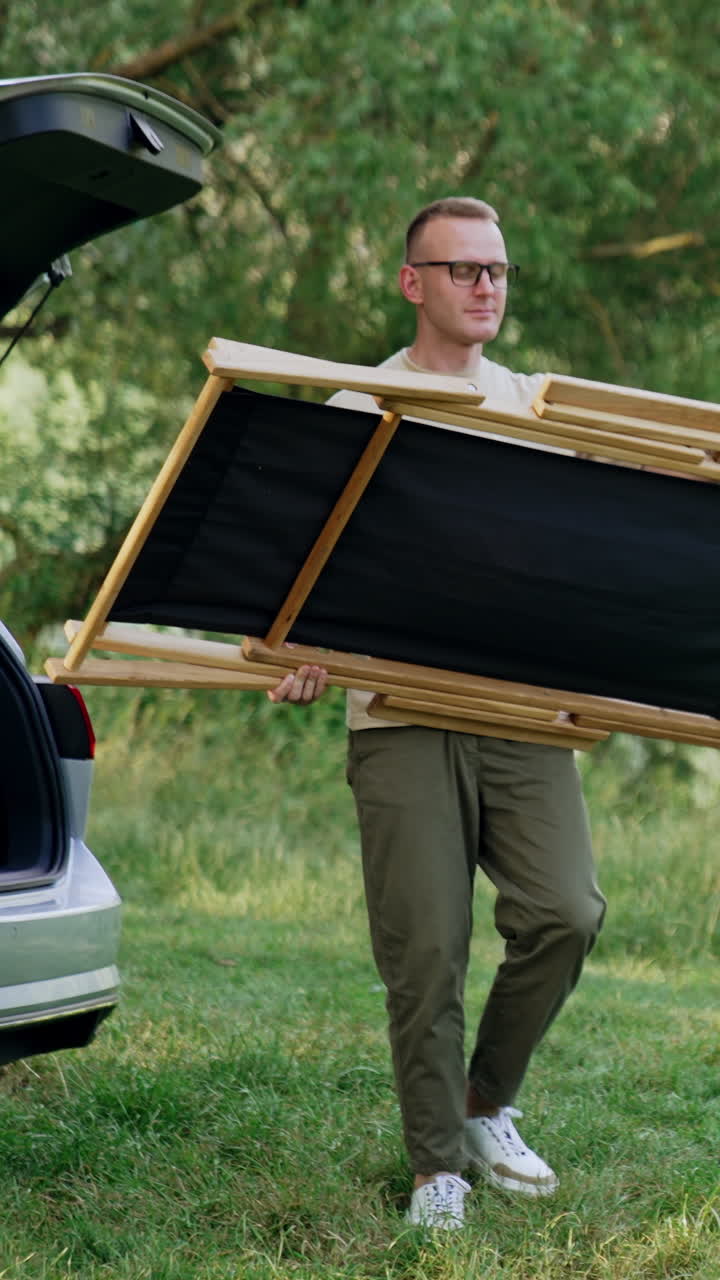 Man comes up to the car trunk. Man takes out the folding chair and puts it on the grass. Rest in nature. Vertical video