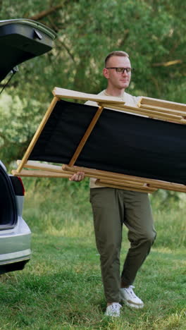 man comes up to the car trunk. man takes out the folding chair and puts it on the grass. rest in nature. vertical video