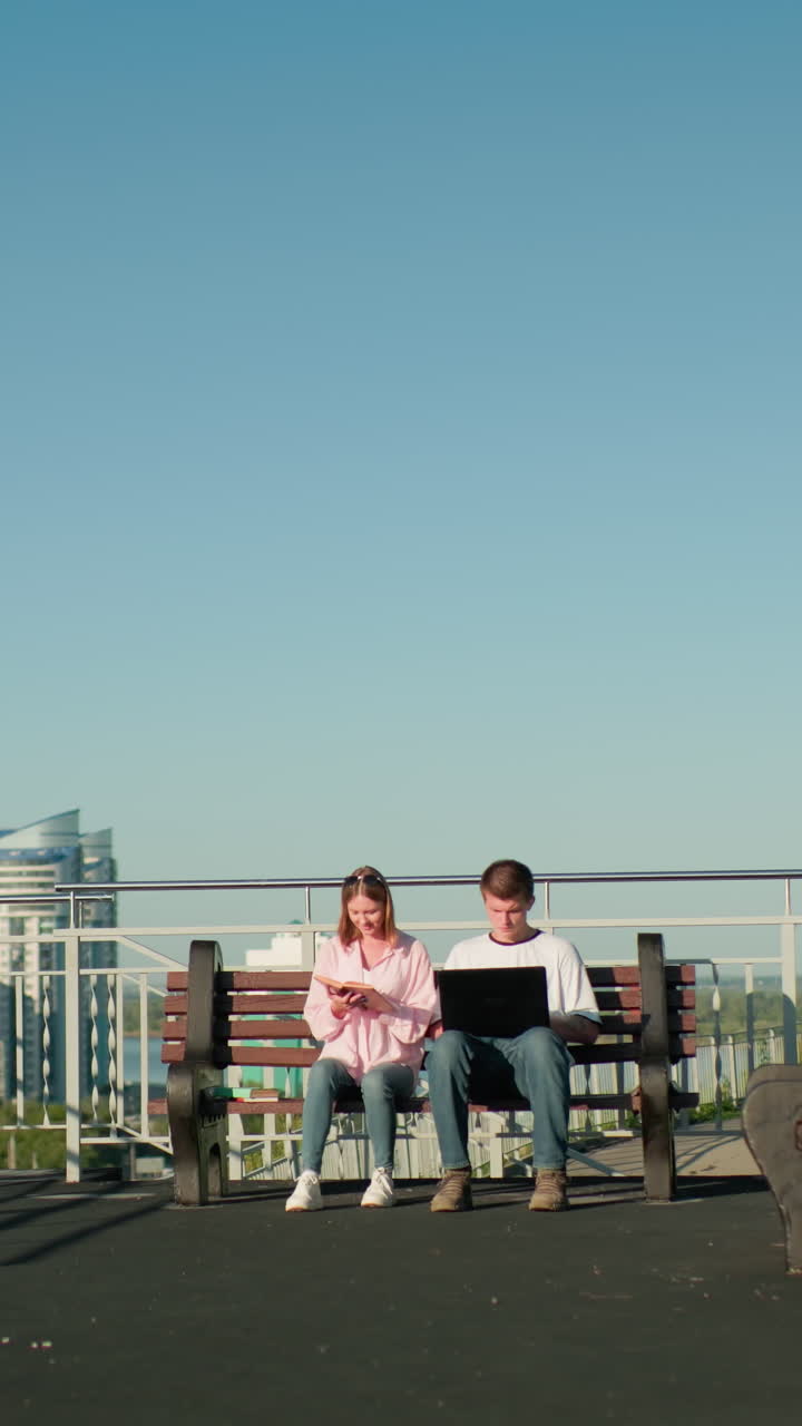 dos estudiantes sentados al aire libre en bancos de madera, un niño escribiendo en una computadora portátil mientras la niña señala su libro, rodeados de paisaje urbano, barandillas, vegetación y cielo azul claro en un día soleado y brillante