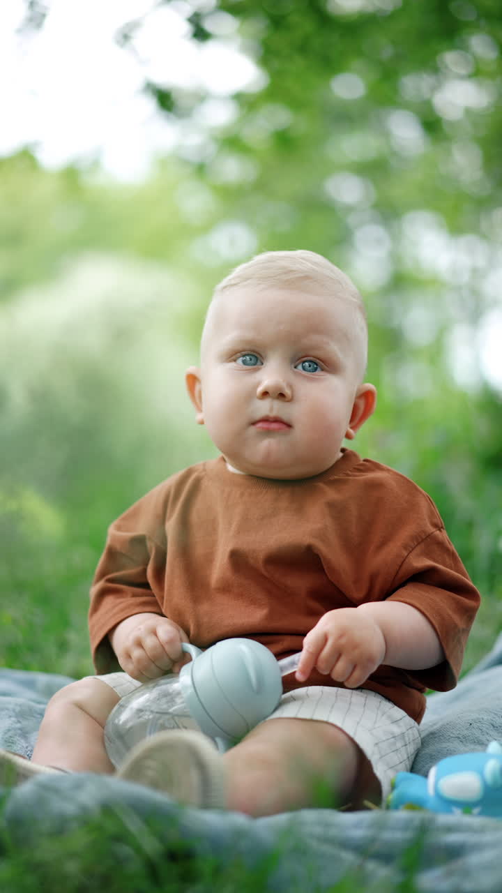 Lovely peaceful blond baby boy in brown t-shirt sits outdoors. Cute toddler holds a water bottle and looks at camera. Blurred backdrop. Vertical video.