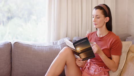 woman relaxing on sofa, reading book and enjoying coffee by window