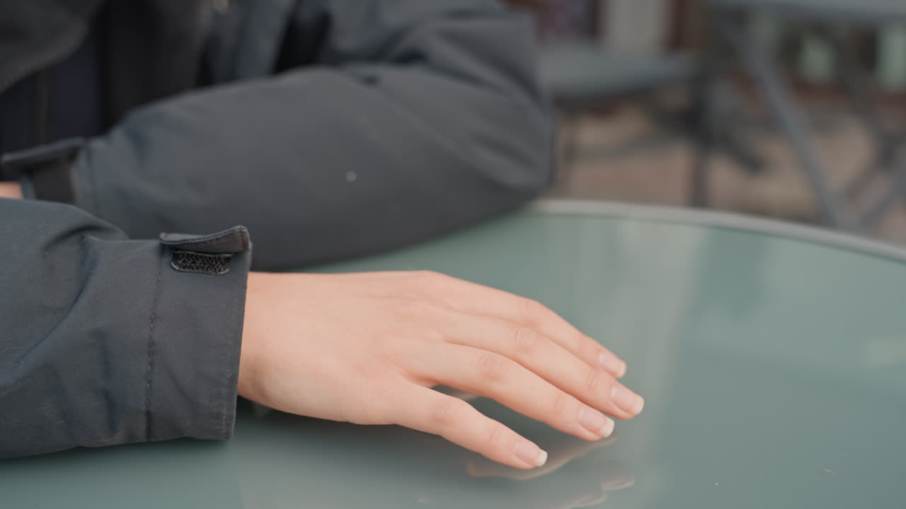 Persons hand resting on glass table, jacket sleeve visible, soft reflection on surface, relaxed fingers, outdoor cafe patio, muted tones, shallow depth of field, waiting mood, subtle fabric texture