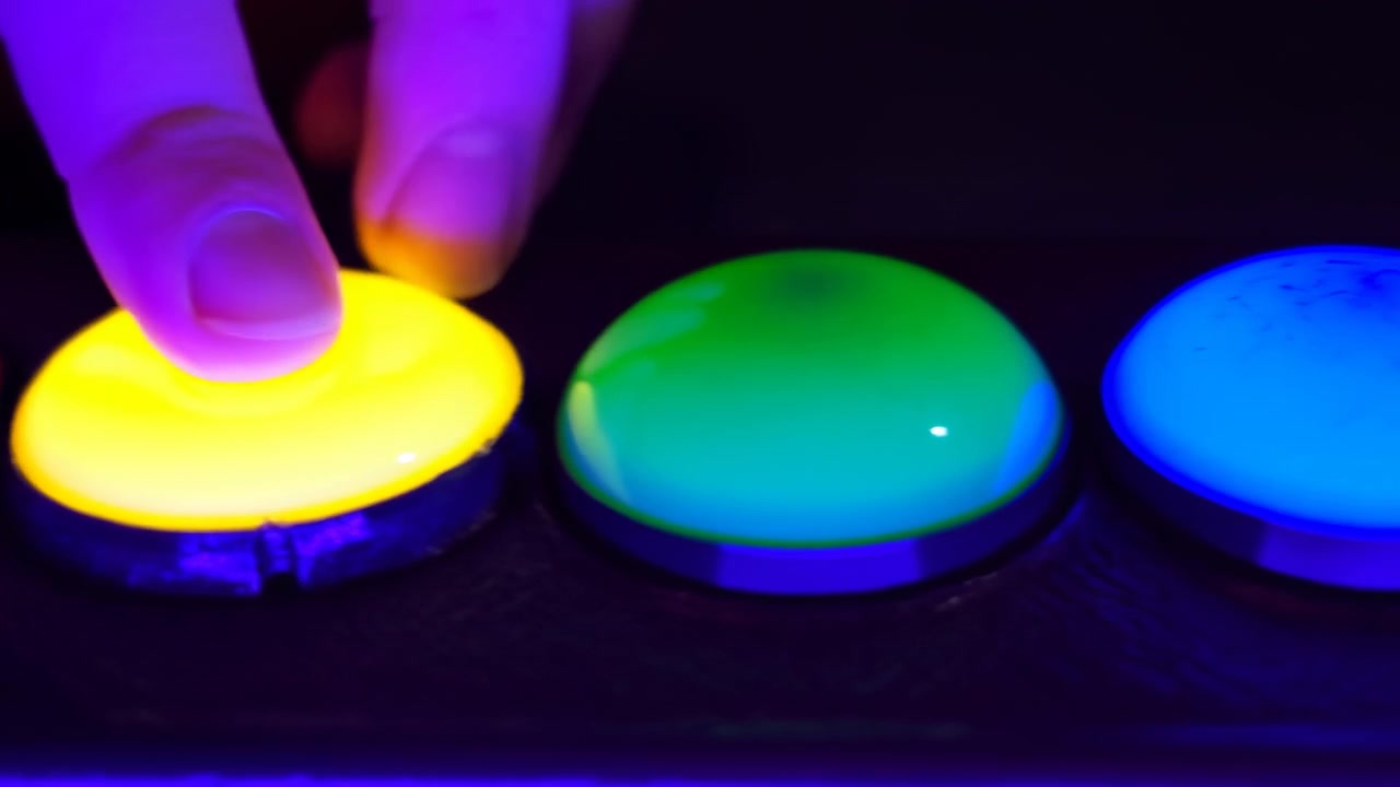 Primo piano di una fila interattiva di pulsanti RGB con un polpastrello che preme cupole lucide in un ambiente scuro e a bassa luminosità, mostrando sfumature di colore luminose su superfici rosse, verdi, blu e giallo caldo.