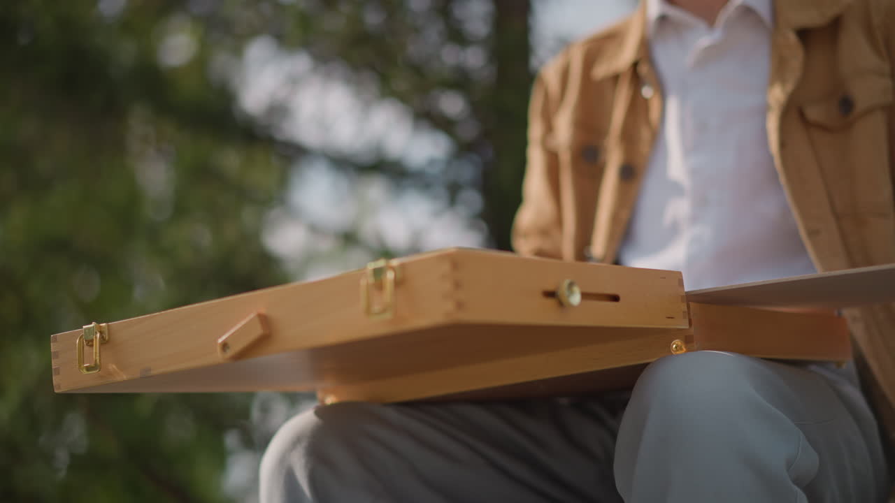 cavalletto portatile apribile sulle gambe, mani che sganciano i fermi e sollevano il coperchio in legno, luce solare che filtra tra gli alberi, momento preparatorio intenso per una sessione di pittura all'aperto, rivelazione di attrezzatura compatta