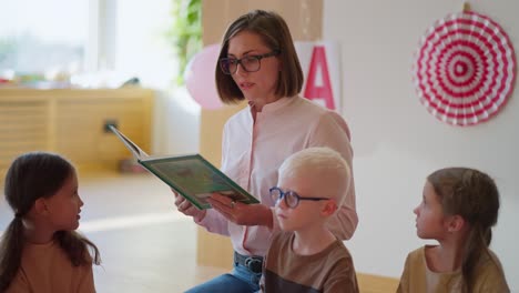 A-blonde-woman-with-a-bob-hairstyle-in-glasses-and-a-white-and-pink-shirt-reads-a-green-book-for-students-in-a-club-for-preparing-children-for-school