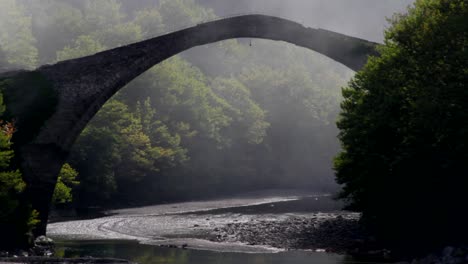 fog surrounding an arched stone bridge in aoos river in greece