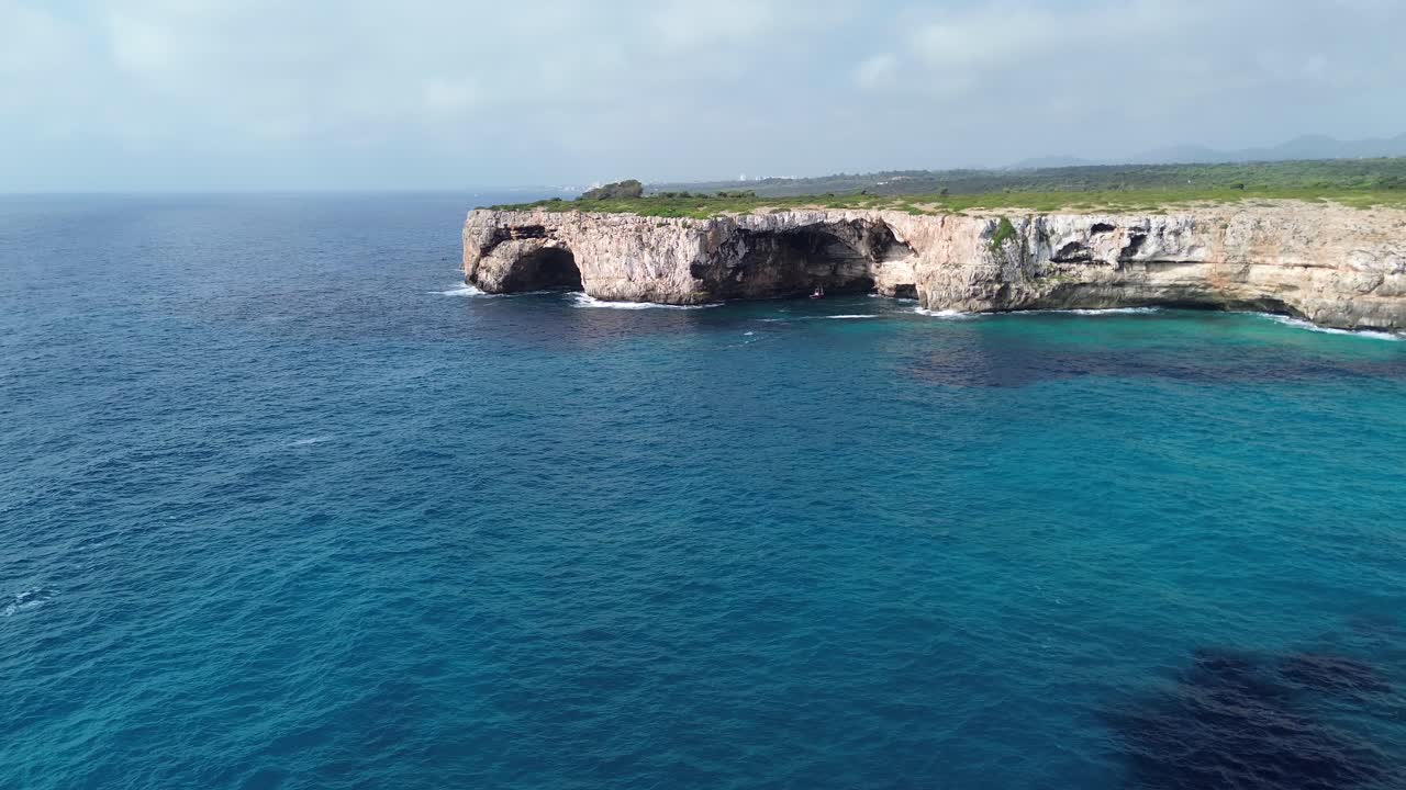 Cinematic 4K drone footage flying horizontally toward two large sea caves along Mallorca’s rugged coastline. Captured on a sunny day in June 2025 with clear blue water, rocky cliffs, and green bushes