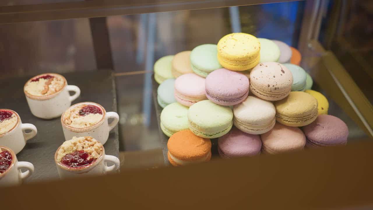 Close up of assorted colorful macarons arranged in pyramid shape beside small dessert cups with berry toppings and crumbles in store display case under warm lighting