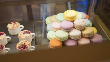 close up of assorted colorful macarons arranged in pyramid shape beside small dessert cups with berry toppings and crumbles in store display case under warm lighting