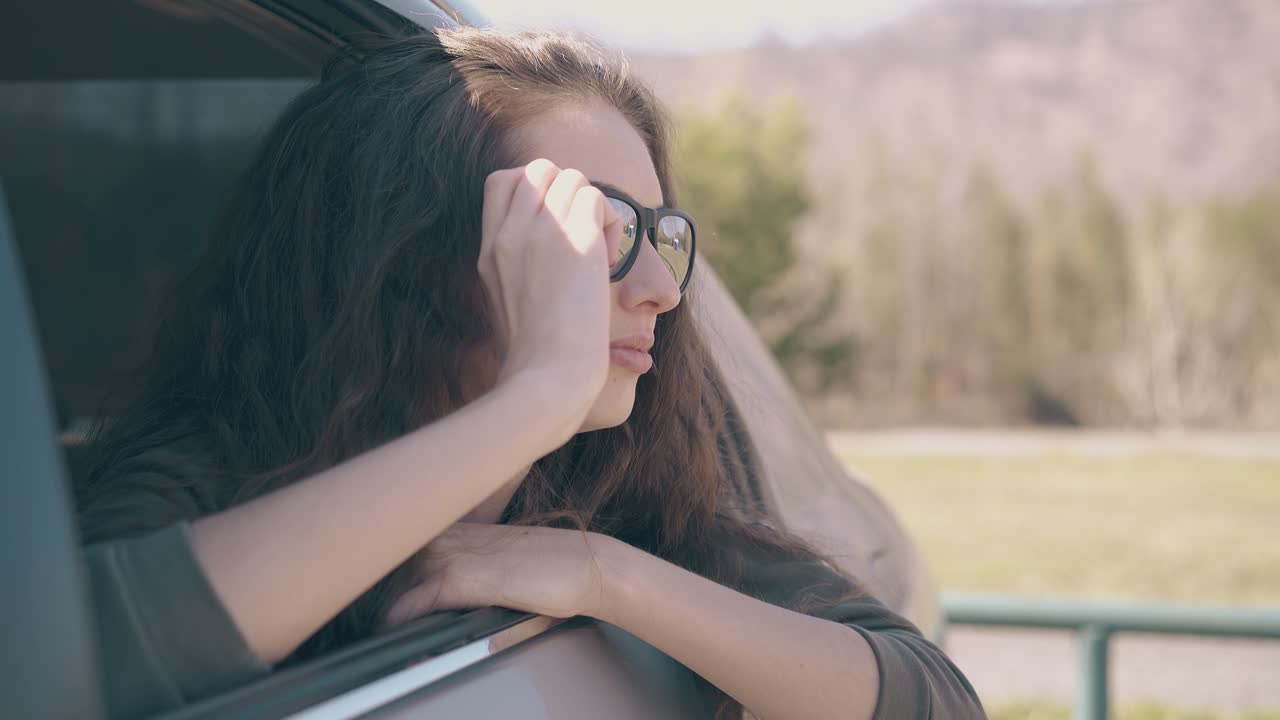 어두운 머리카락을 가진 우아한 여성이 언덕에서 열린 자동차 창문  ⁇ 에 앉아 있습니다.