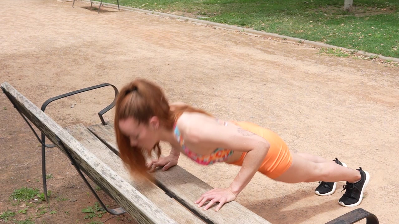 Una donna impegnata in una routine di fitness all'aperto esegue flessioni inclinate su una panchina del parco.
