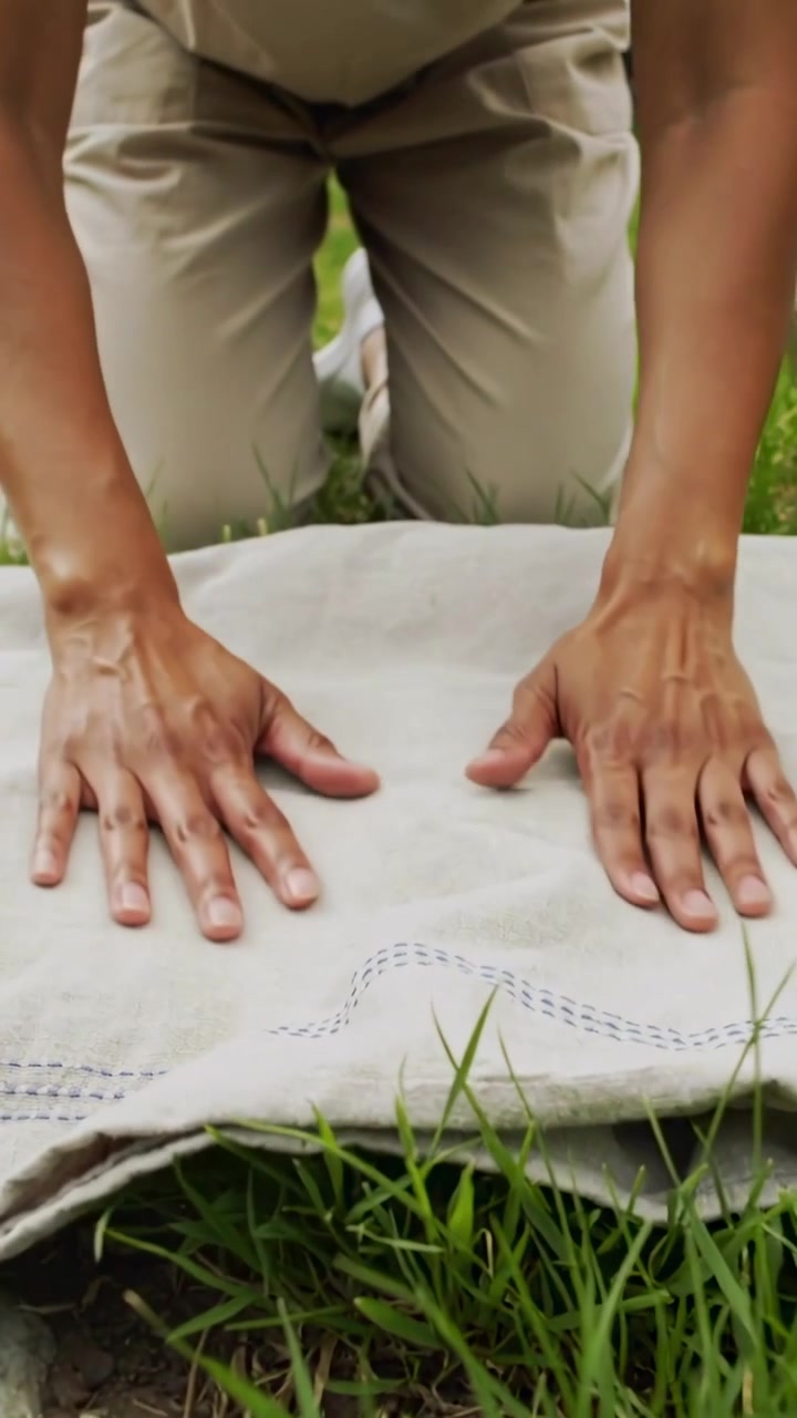 Video verticale: Adulto inginocchiato in giardino, con pantaloni beige, che si china per premere e lisciare un panno