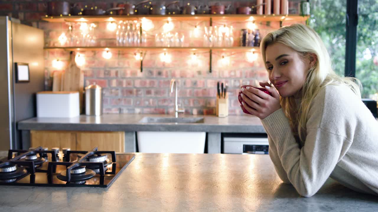 Woman lifting red ceramic mug, sipping coffee while smiling toward camera in kitchen, copy space