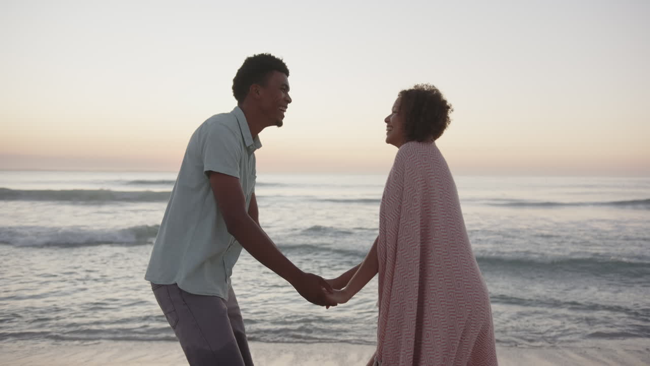 Biracial couple holds hands on the beach at sunset, with waves in the background