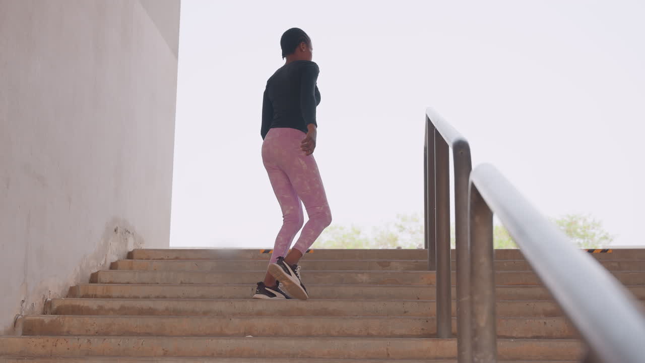 Mulher correndo escadas de concreto, entusiasta de fitness matinal sobe escadaria com a mão no corrimão, vestindo calças rosa e top preto, passo focado e postura confiante, arquitetura minimalista