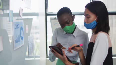 Diverse-female-office-colleagues-wearing-face-masks-discussing-over-digital-tablet-at-modern-office