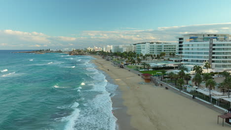 Drone-View-of-Turquoise-Sea-Water-Waves-Rolling-on-Sandy-Beach-in-Protaras-with-Beachfront-Hotel-Resorts-at-Sunset,-Cyprus