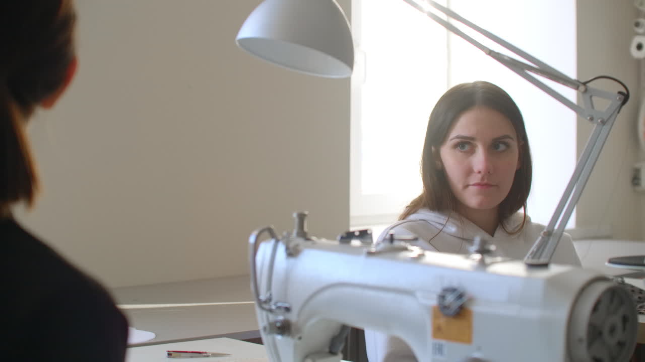American seamstress with measuring tape over her neck applying threads to discussing it with cheerful Caucasian female colleague while cooperating in design studio.