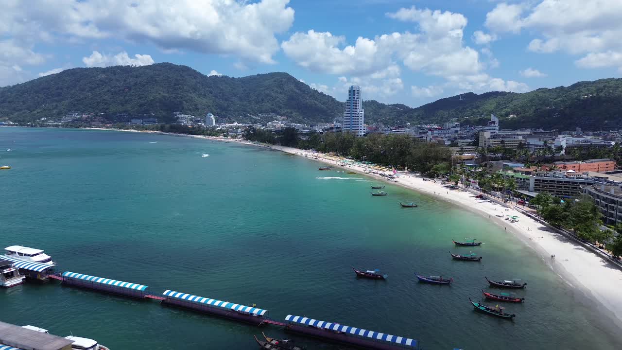 Toma de dron de la playa de Patong con múltiples barcos alrededor de un muelle flotante a lo largo de una concurrida costa tropical con hoteles y aguas cristalinas en Phuket, Tailandia, un popular destino turístico.