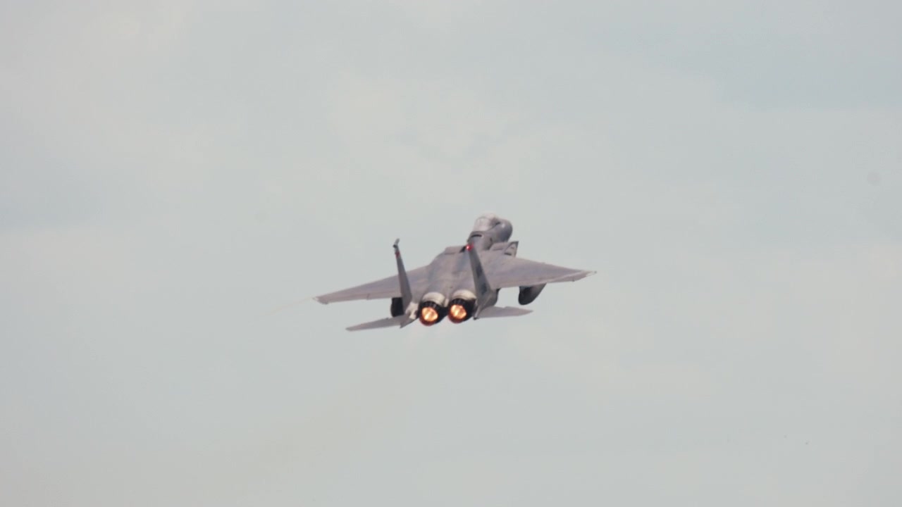 A military fighter jet banks through an overcast sky with glowing afterburners.