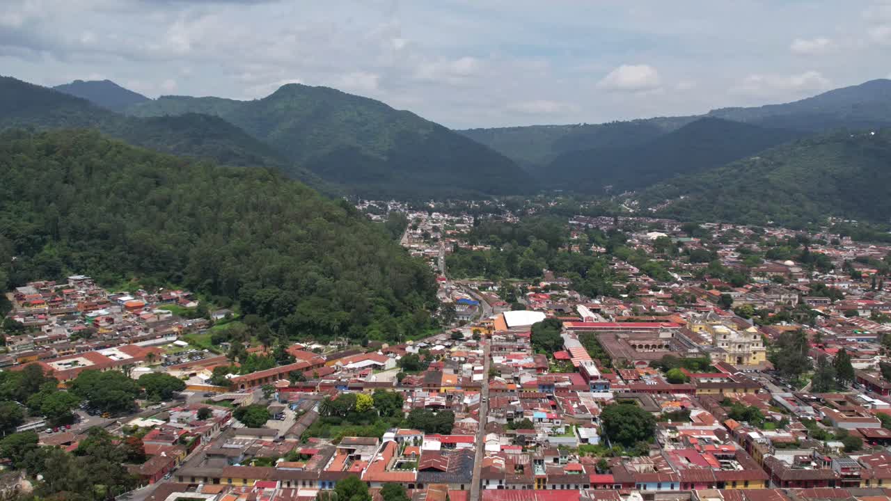 Hyperlapse of Antigua, Guatemala panning through town showing colorful red rooftops, green trees, traffic in streets, and revealing volcano Volcan De Agua on a cloudy bright day.